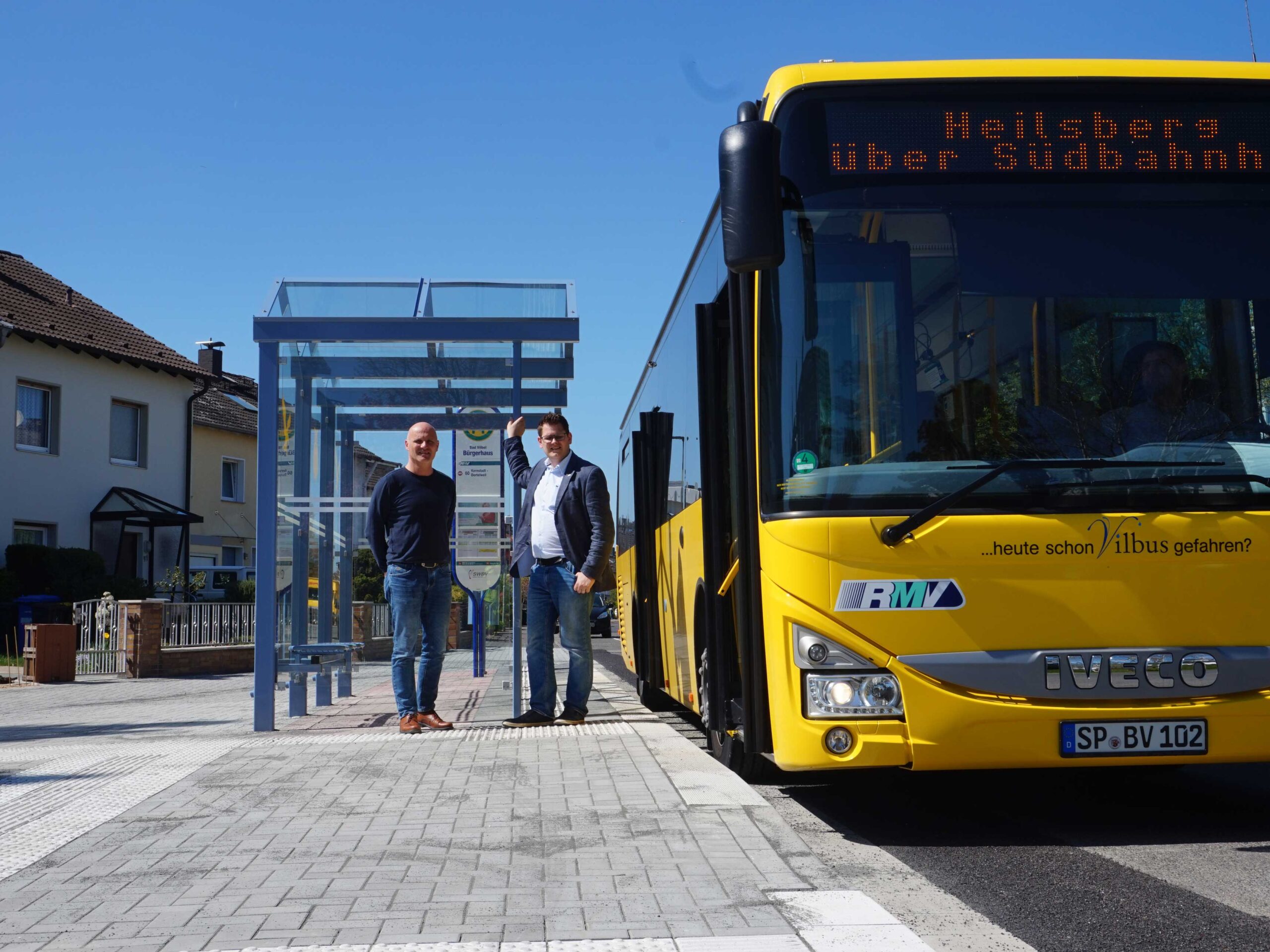 2 Personen stehen an einer Haltestelle, wo ein gelber Vilbus steht